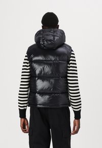 Vesta puffer lucioasă, de culoare neagră, cu un glugă mare, având un design matlasat. Modelul poartă un pulover cu dungi negre și albe dedesubt.