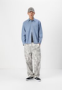 Lyseblå knap-op skjorte, grå mønstrede bukser og sorte sneakers. Skjorten har en brystlomme; bukserne er løstsiddende med et detaljeret print.