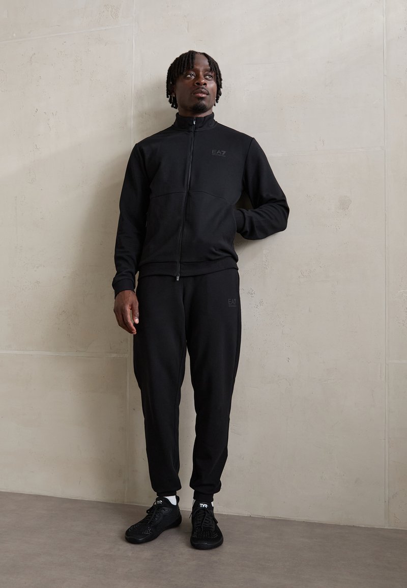 Schwarzer Sportanzug aus weichem Material, bestehend aus einer Zip-Jacke und passenden Hosen mit elastischen Bündchen, akzentuiert durch ein Logo.