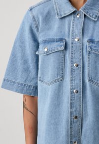 Lyseblå denim skjorte med korte ermer, to brystlommer, sølvfargede trykknapper og en myk tekstur. Har synlige sømdetaljer.