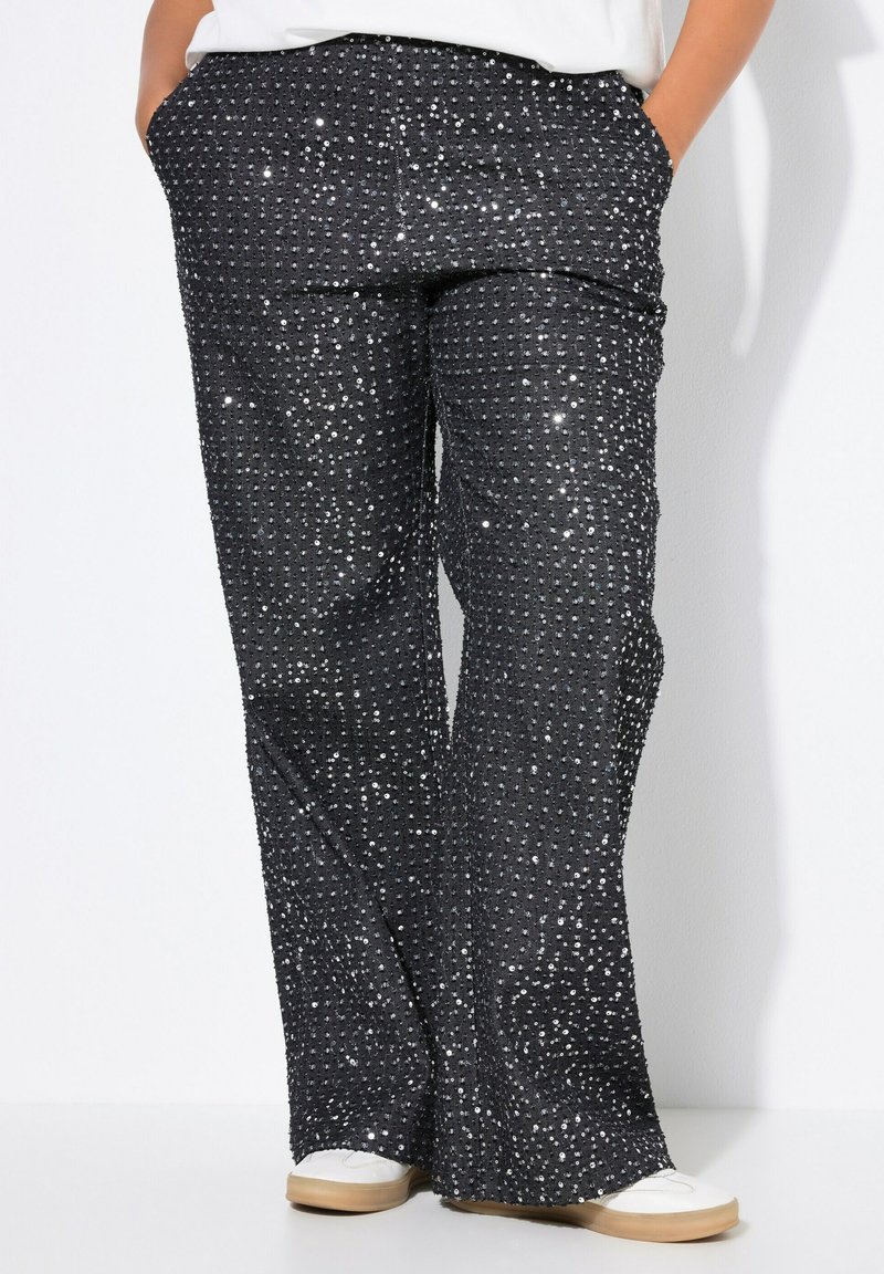Pantalones de lentejuelas negros con un diseño de pierna ancha, que presentan una superficie texturizada de pequeñas lentejuelas brillantes y bolsillos laterales. Parte superior y zapatos blancos minimalistas.