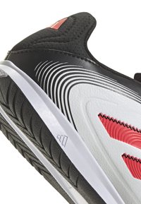 Zapatilla deportiva en blanco y negro con detalles en rojo, que presenta un upper texturizado, diseño contorneado, rayas dinámicas y suela de goma con patrón de tracción.
