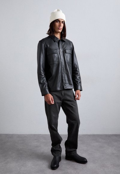 Black leather shirt jacket with two chest pockets, worn over dark jeans and black boots. Model wearing a cream beanie. Neutral backdrop.