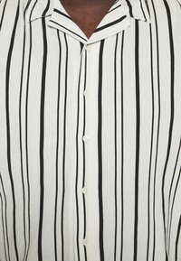 Mies, joka on pukeutunut valkoiseen nappipaitaan, jossa on mustia pystysuoria raitoja ja avoin kaulus, joka paljastaa rintaihoa.