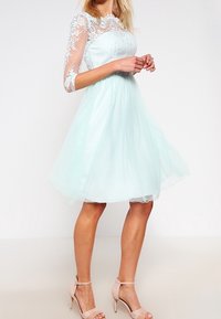 Robe vert menthe avec une jupe en tulle, des manches trois-quarts en dentelle et un corsage brodé. Portée avec des chaussures à talons hauts beiges à lanières.