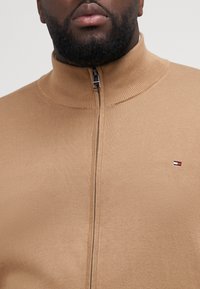 Beżowy sweter zapinany na zamek z wysokim kołnierzem, tekstura w prążki oraz mała naszywka z logo w czerwono-biało-niebieskich kolorach na klatce piersiowej.