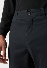Pantalons en coton noir avec une coupe droite, fermeture à bouton sur le devant et détails de couture visibles. Le matériau a une texture lisse.