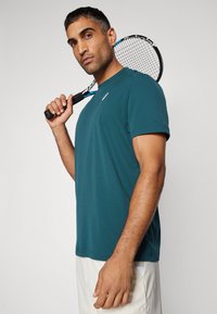 Teal athletic t-skjorte med korte ermer, med en liten logo på brystet. Modellen holder en tennisracket, som viser en strømlinjeformet passform.