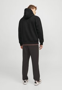 Schwarzer Kapuzenpullover mit Kängurutasche, gerippten Bündchen und Saum, getragen über einem braunen Shirt, kombiniert mit lockeren schwarzen Hosen und Sneakers.