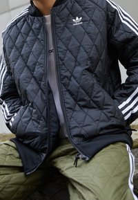 Svart quiltad bomberjacka med vitt Adidas-märke och tre ränder på ärmarna, kompletterad med olivgröna byxor med diamantmönster.