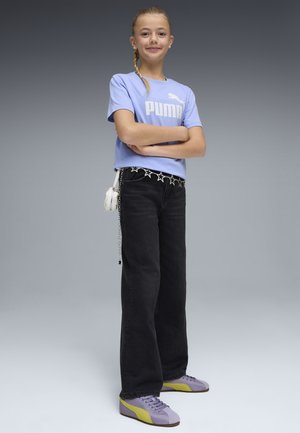 Młoda dziewczyna stojąca z założonymi rękami, ubrana w liliową koszulkę Puma, czarne jeansy, pasek z łańcuszkiem w kształcie gwiazd, małą białą torebkę oraz liliowo-żółte trampki.