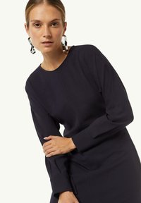 Marineblaues langärmliges Kleid mit Rundhalsausschnitt und seitlichen Nahtdetails. Hat eine glatte Textur, eine figurbetonte Silhouette und Akzente mit silbernen Ohrringen.