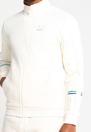 Mann trägt eine weiße Puma-Jacke mit Reißverschluss und grünen sowie blauen Streifen an den Ärmeln, Hände in den Taschen, vor einfachem Hintergrund.