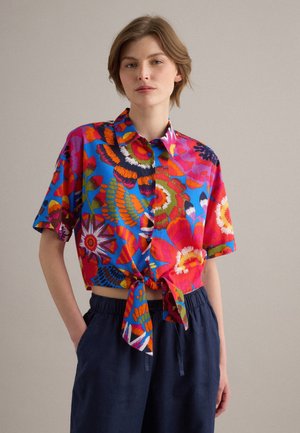 Joven con pelo corto que lleva una camisa colorida de flores de manga corta abotonada, atada a la cintura, y pantalones oscuros y sueltos, de pie relajado.