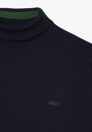 Navy coltrui van gebreid materiaal met een geribde groene kraag en een subtiele, geborduurde logo op de borst. Zachte textuur en aangesloten pasvorm.