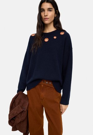 Kobieta w granatowym swetrze z okrągłymi wycięciami i pomarańczowo-brązowych spodniach, trzymająca brązową kurtkę w lewej ręce.