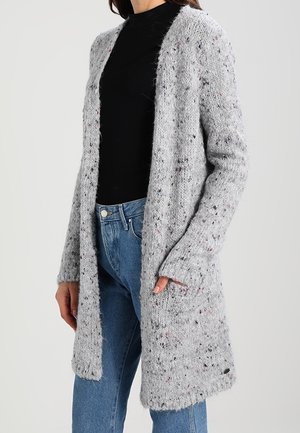 Frau trägt eine lange, grau gesprenkelte Strickjacke über einem schwarzen Oberteil und blauen Jeans, steht mit einer Hand in der Tasche.