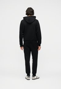 Άνδρας στέκεται γυρισμένος προς τα πίσω, φοράει μαύρη φούτερ με κουκούλα, μαύρο παντελόνι και λευκά αθλητικά παπούτσια με μαύρες λεπτομέρειες σε λευκό φόντο.