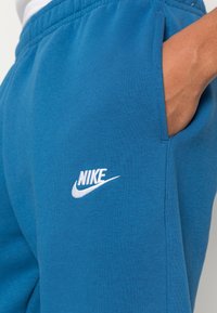 Blå bomullssweatpants med elastisk midja, sidofickor och en vit broderad Nike-logotyp på vänstra benet. Mjuk textur.