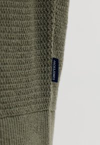 Gros plan d'un tissu en maille vert olive avec un motif texturé et une petite étiquette bleu foncé portant la mention "JACK&JONES" cousue dans la couture.