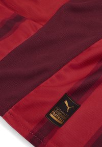 Fotbollströja med en röd och mörkröd färgskala, nätstruktur och en guldtagg med "AUTENTISKT LICENSIERAD FOTBOLL" och Puma-logotyp.