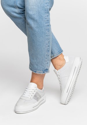 Person wearing light blue cropped jeans and white low-top sneakers, one foot lifted slightly off the ground against plain background.