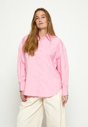 Žena s dlhými vlasmi, oblečená v voľnej ružovej košeli na gombíky a širokých bielych nohaviciach, stojaca pred jednoduchým svetlým pozadím.