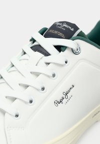 Balta ādas sneaker ar teksturētu virsmu, zaļām detaļām un gumijas zoli. Ir baltas mežģīnes un logotips pusē un mēlītē.