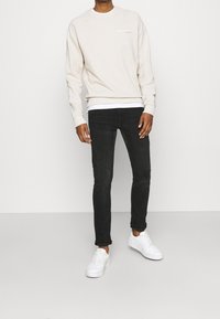 Man som bär en beige sweatshirt, svarta slim-fit jeans och vita sneakers, står på ett enfärgat ljust grått golv mot en vit vägg.