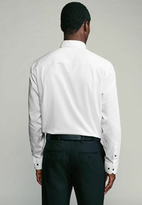Chemise blanche à manches longues avec un col structuré, texture lisse et accents de poignets noirs, associée à un pantalon sombre avec des détails de poche arrière.