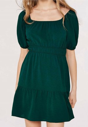 Femme portant une robe courte vert foncé avec manches bouffantes, encolure carrée, et taille froncée, debout devant un fond neutre.