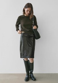 Dunkelgrüner Pullover, glänzender schwarzer Leder-Bleistiftrock, lange schwarze Gummistiefel und eine strukturierte schwarze Schultertasche.