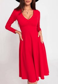 Robe rouge à manches longues avec un corsage ajusté et une jupe évasée. Caractérisée par un décolleté en V plongeant et un détail froncé au niveau de la poitrine, confectionnée en tissu lisse.
