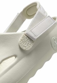 Białe slip-on buty z teksturowanego materiału gumowego, z regulowanymi paskami z tkaniny i okrągłym logo z graficznym emblematem.