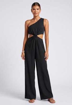 Vrouw draagt een zwarte jumpsuit met één schouder, zijopeningen en brede pijpen, staand tegen een effen lichtgrijze achtergrond.