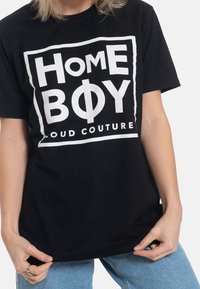 Svart bomullströja med en djärv vit grafisk tryck som säger "HOME BOY" och "LOUD COUTURE" inom en rektangulär ram.