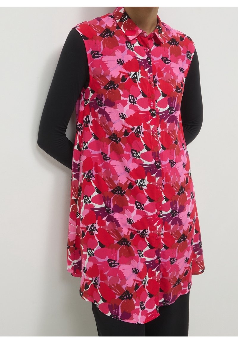 Personne portant une tunique sans manches boutonnée avec un motif floral rouge, rose et violet sur un haut à manches longues noir et un pantalon noir.