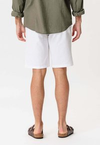 Mand iført hvide shorts til knæene, olivengrøn skjorte med lange ærmer der er optrukket, og brune sandaler med åben hæl, stående og vendt væk.