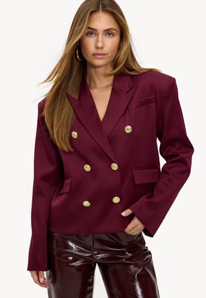 Femme aux longs cheveux portant un blazer bordeaux à double boutonnage avec des boutons dorés et un pantalon brillant bordeaux foncé, posant avec une main dans la poche.