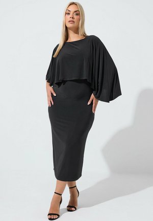 Frau mit langen blonden Haaren, die ein schwarzes Midikleid mit weiten, fließenden Ärmeln und schwarzen High-Heel-Sandalen trägt, steht vor einem schlichten Hintergrund.