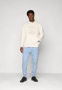 Lichtbeige hoodie met een grafisch logo, gecombineerd met lichtblauwe cargobroek van sweatstof en zwarte sneakers. Materiaal lijkt zacht; ontwerp is casual.