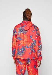 Chaqueta roja con patrón floral azul y amarillo, capucha, puños elásticos y dobladillo. Material ligero con un corte relajado, acompañado de pantalones a juego.