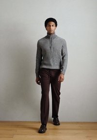 Camisola cinzenta canelada com decote com fecho éclair, combinada com calças castanho escuro feitas à medida e sapatos castanhos, em pé sobre um chão de madeira contra uma parede clara.