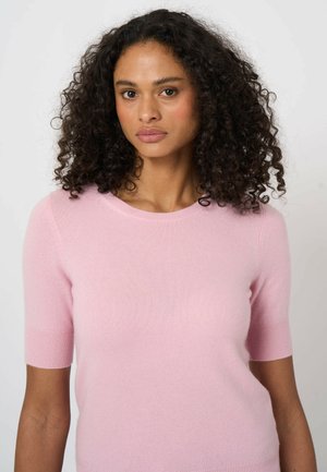 Kobieta o kręconych ciemnych włosach, ubrana w jasno różowy krótki sweter z krótkim rękawem, patrząca prosto w obiektyw na jednolitym tle.