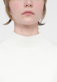Suéter branco de gola alta com uma textura canelada, apresentando um design ajustado. Tecido suave, estilo minimalista e sem padrões proeminentes.