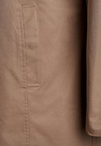 Jasnobrązowa kurtka o gładkiej, matowej fakturze. Posiada boczną kieszeń i przeszycia jako akcenty, z prostym krojem i długimi rękawami.