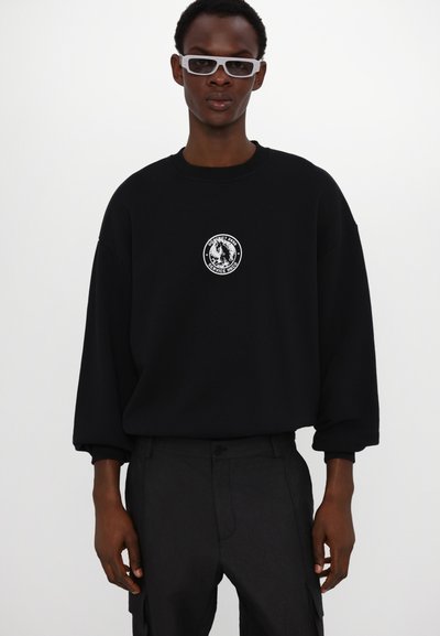 Sweatshirt noir surdimensionné avec un logo graphique circulaire blanc sur la poitrine, associé à un pantalon foncé et des lunettes rectangulaires blanches.