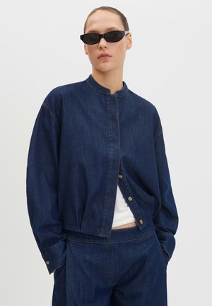 Frau trägt dunkelblaue Jeansjacke und Hose mit den Händen in den Taschen, kombiniert mit kleinen schwarzen Sonnenbrillen und einem weißen Top darunter.