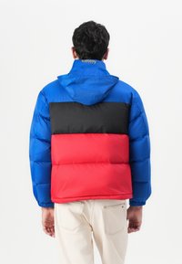 Padded jacket s modrou hornou časťou, čiernou strednou časťou a červenou dolnou časťou. Obsahuje kapucňu a elastické manžety, vyrobený z hladkého, lesklého materiálu.
