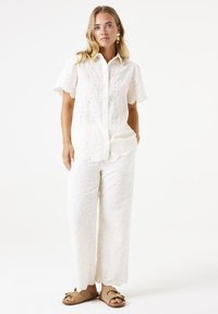 Camisa blanca de algodón con botones y mangas cortas, con intrincados bordados florales y detalles en corte, combinada con pantalones de pierna ancha a juego.